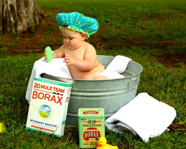 Hatted Infant In A Wash Tub, Holding A Bar Of Green Soap, A Box And Tin Of Borax Nearby