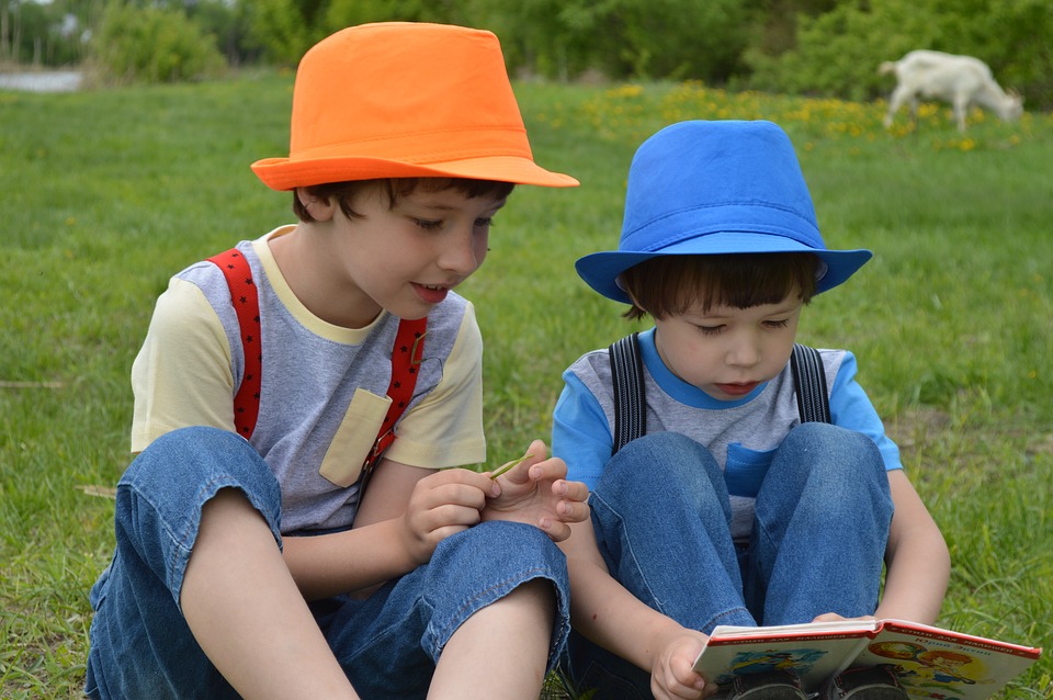 Brothers Sitting On Green Grass, Wearing Colorful Orange And Blue Hats, Reading A Book