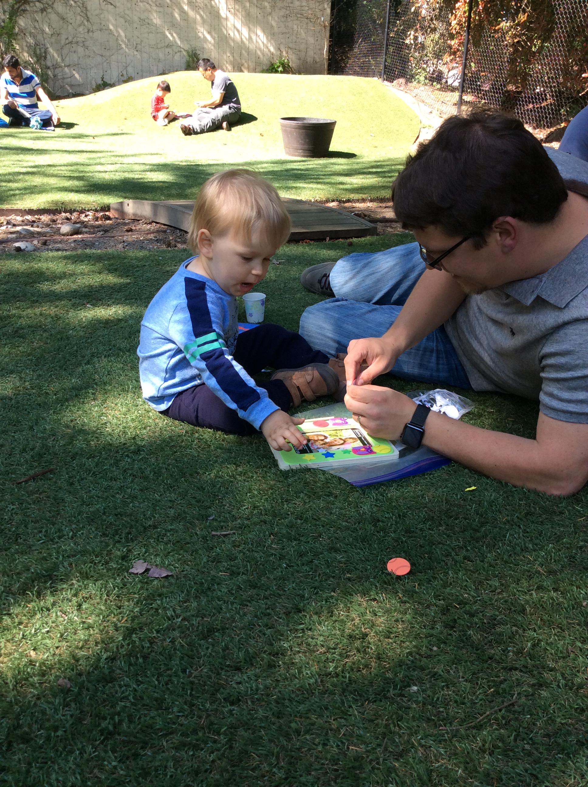 A loving father and his little boy are having fun sitting in the grass playing with stickers at the park.